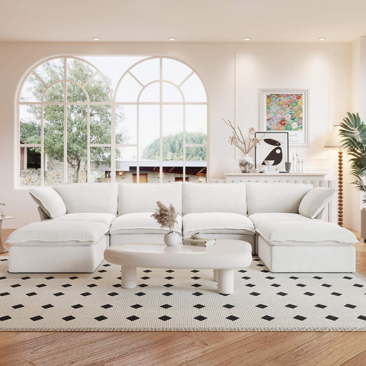 A white U-shaped linen sectional sofa with a storage ottoman situated in a bright room with large windows, alongside a low coffee table with items on it.