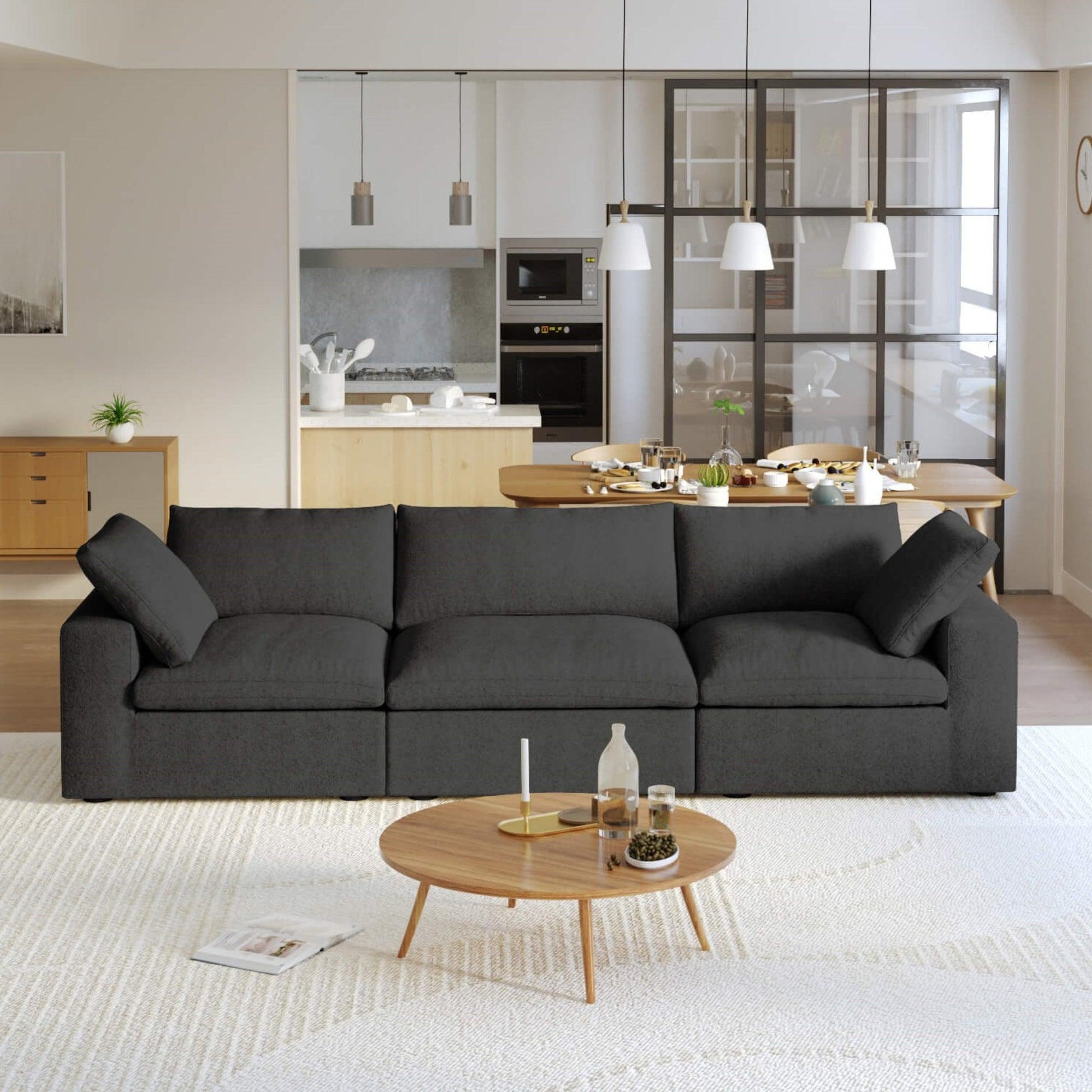 A modern black 3-seater sectional sofa in a living room setting with a round wooden coffee table in front and a kitchen area in the background.