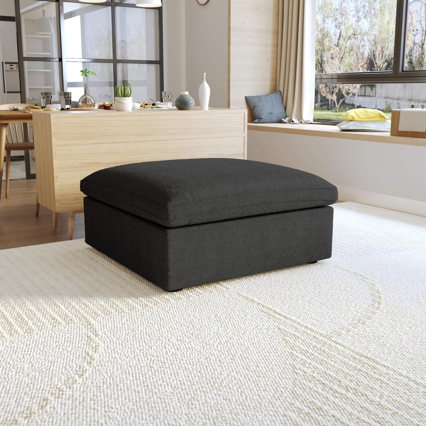 A black, down-filled comfort storage ottoman, placed in a bright living room setting.
