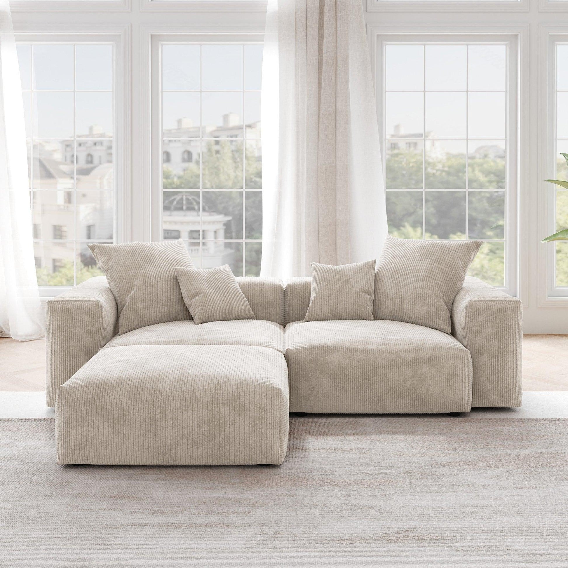 A beige corduroy sectional modular sofa with detachable armrests and removable covers, featuring a chaise lounge configuration placed in a room with large windows.