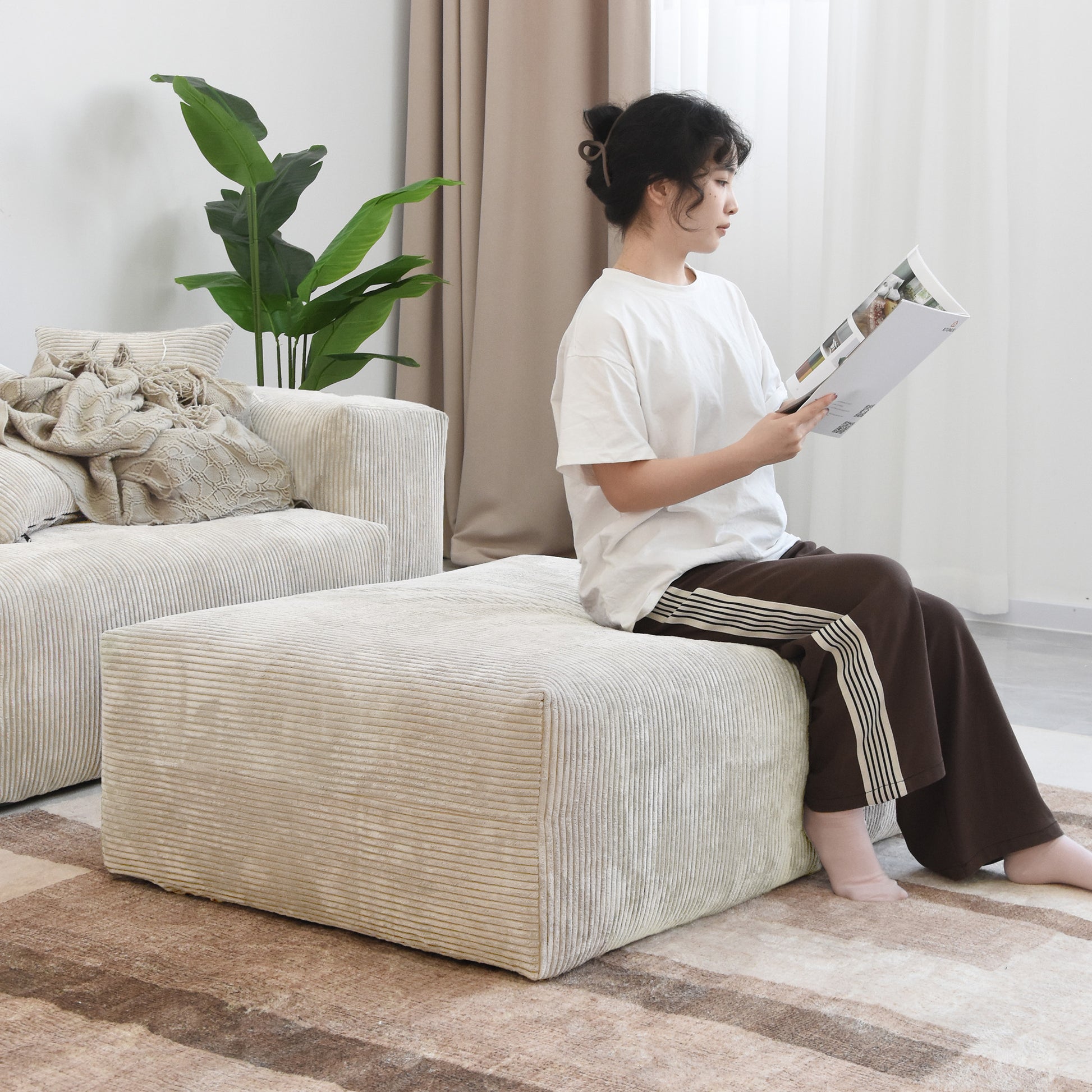 Person sitting on a beige corduroy ottoman, reading magazine in a light living room