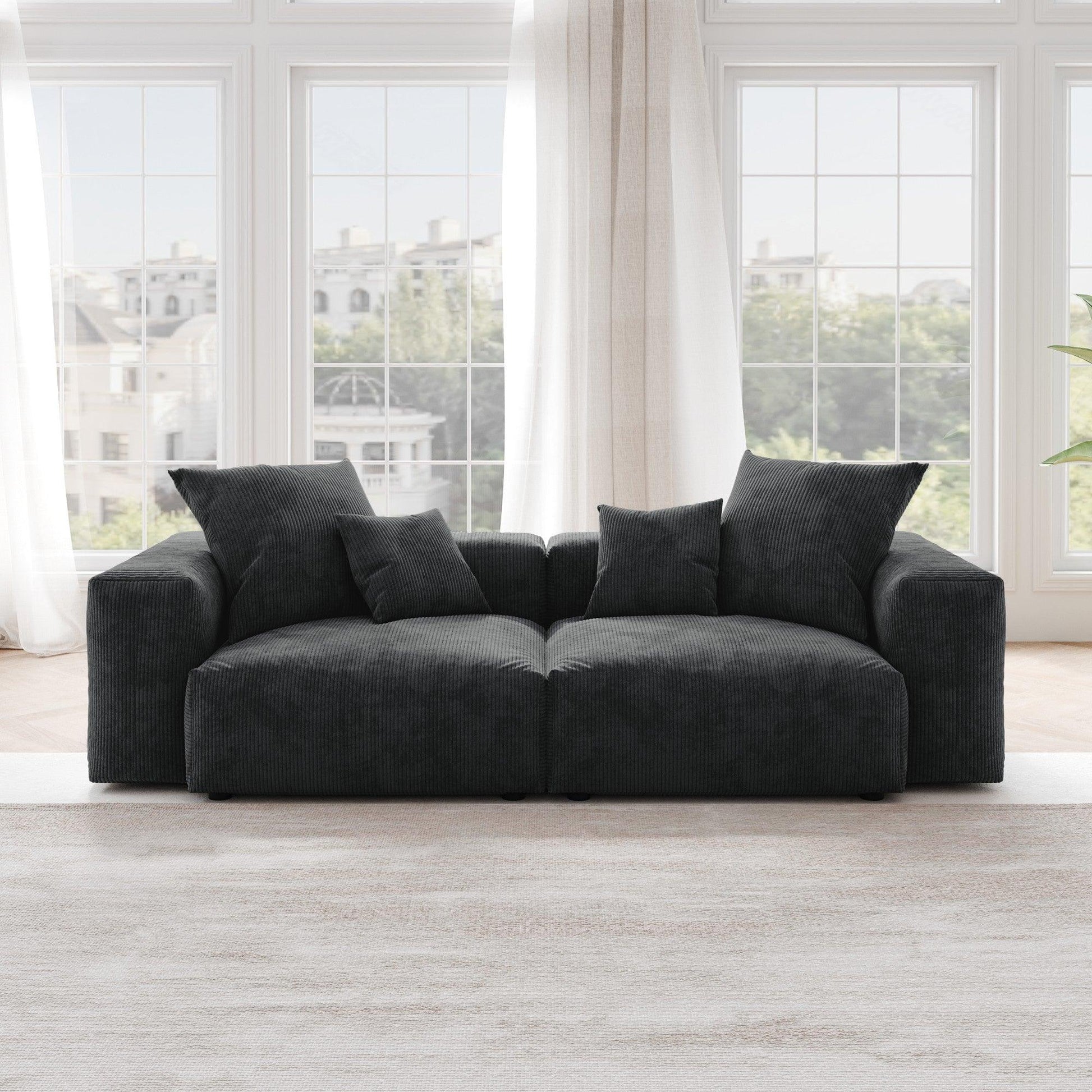 A black corduroy sectional loveseat sofa with detachable armrests and removable covers, positioned in a bright room with large windows.