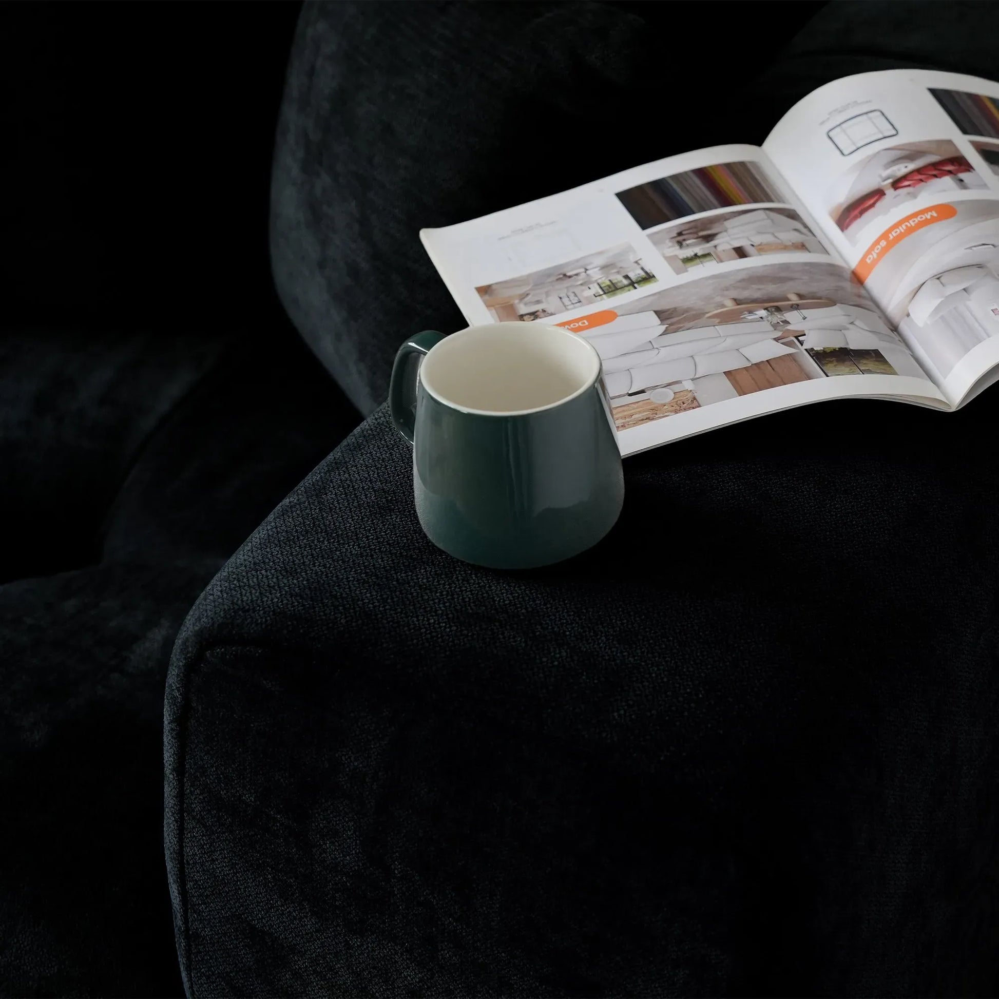 Green mug next to an open magazine on a wide armrest of a black sofa