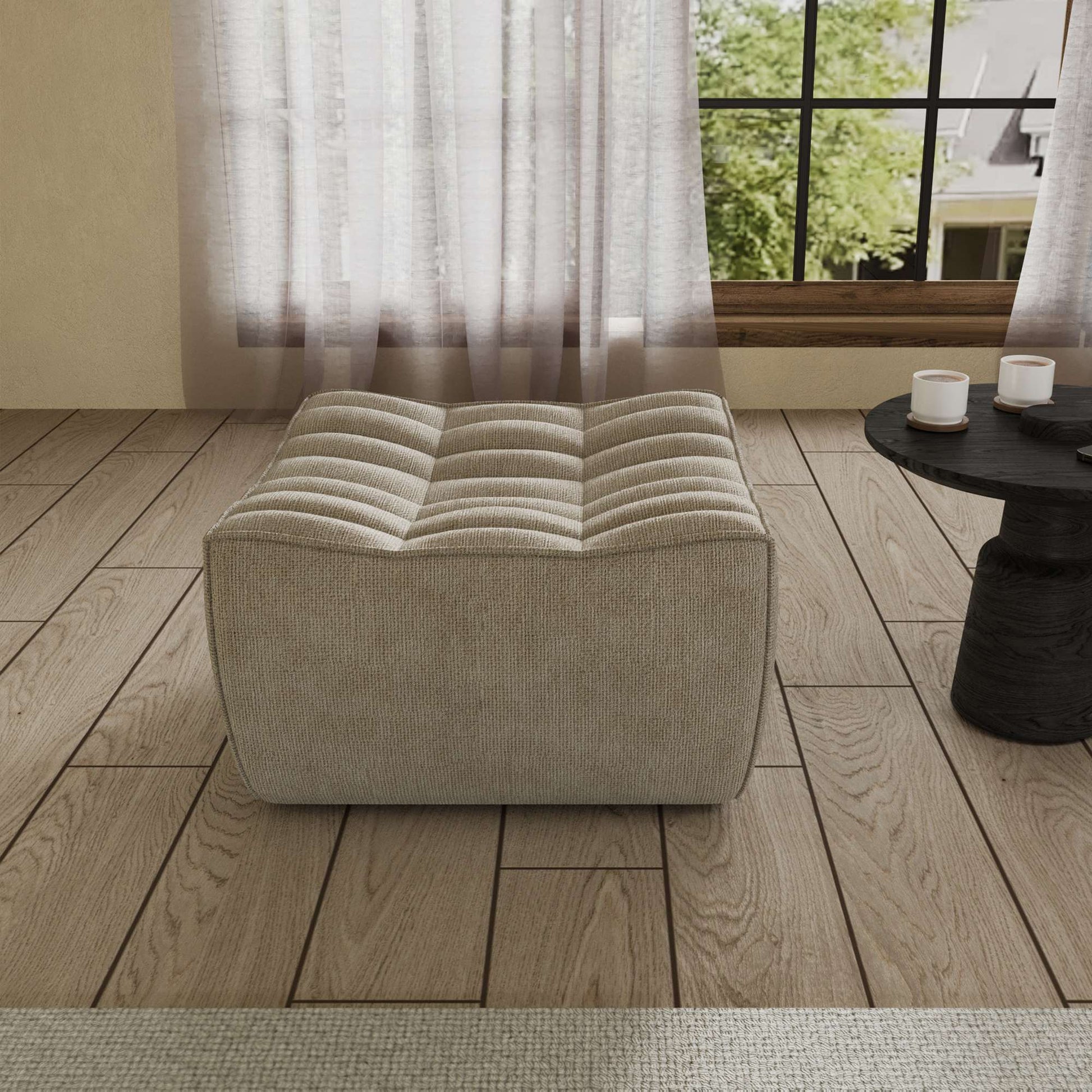 A khaki chenille ottoman with a minimalist design, placed on a wooden floor with a large window in the background.