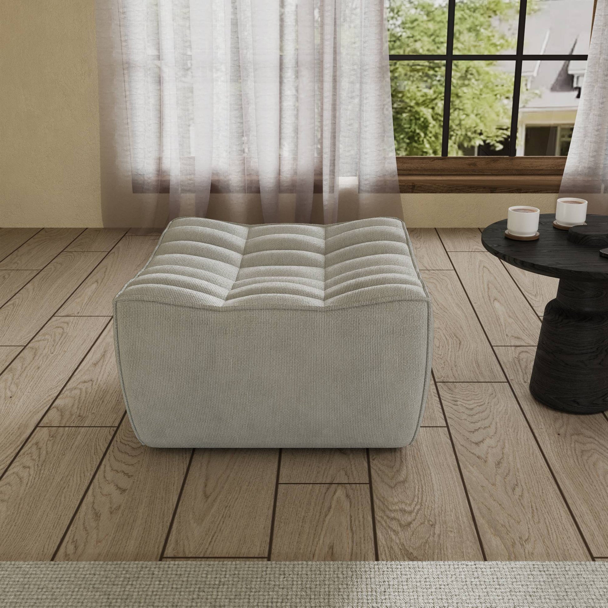 A light gray chenille ottoman with a minimalist design, placed on a wooden floor with a large window in the background.