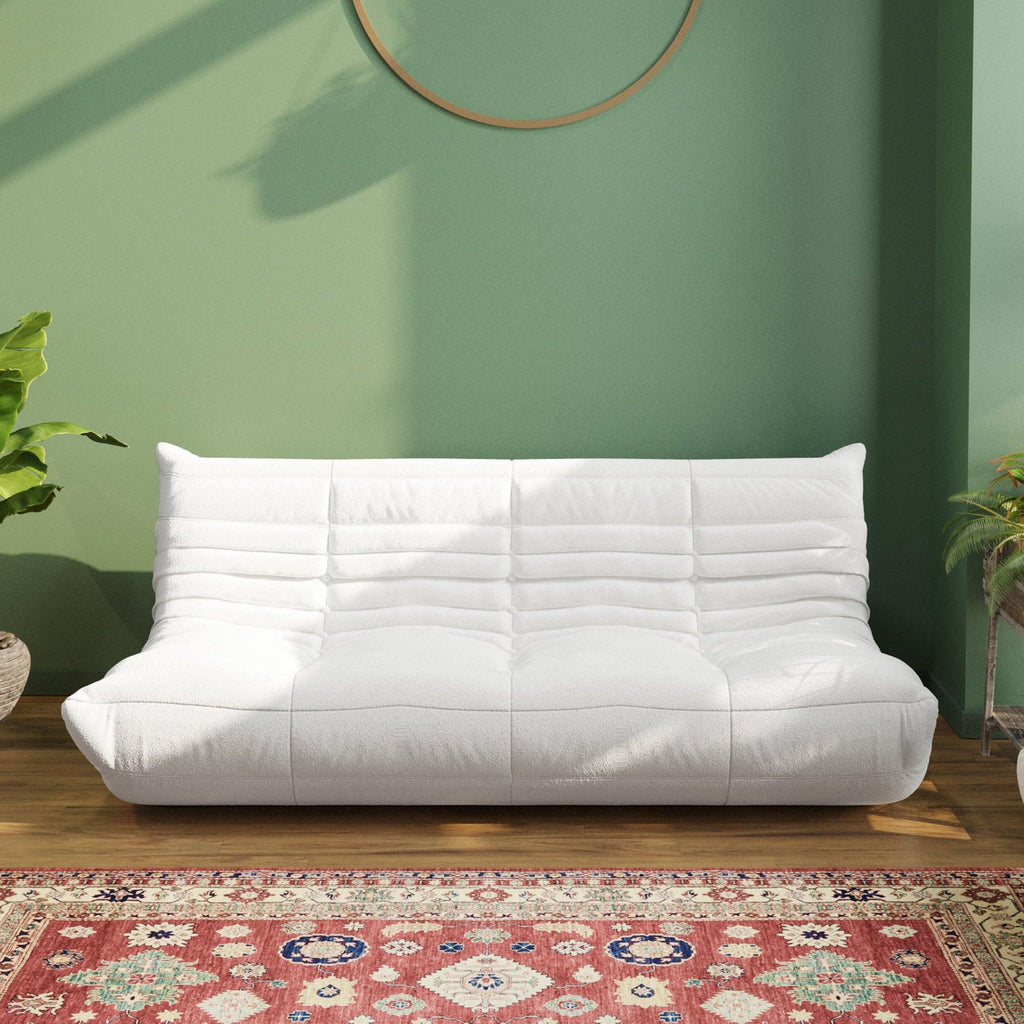 A white, caterpillar-shaped Togo sofa with a plush texture, situated in a room with minimalist decor, on a patterned rug with green wall on the background.