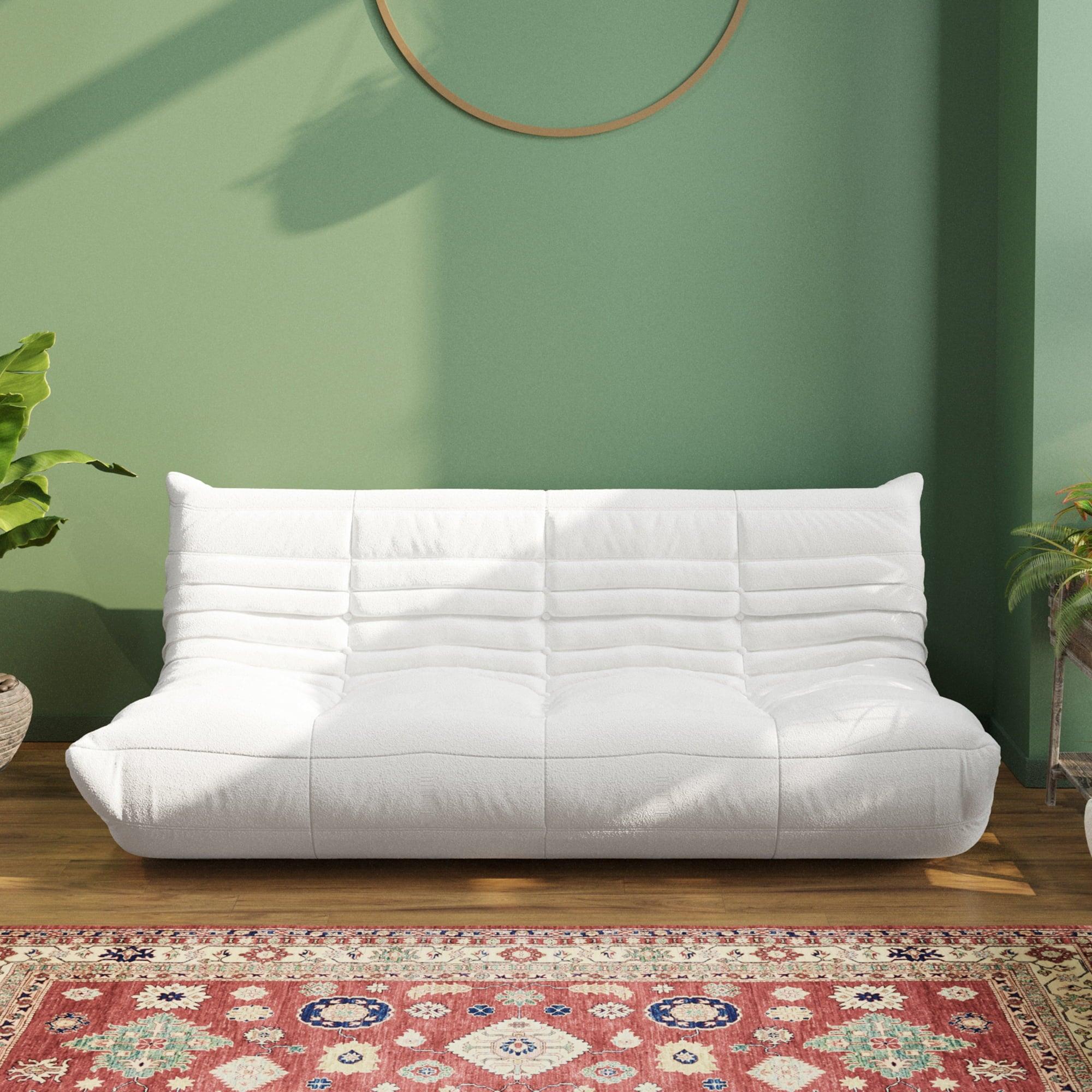 A white, caterpillar-shaped Togo sofa with a plush texture, situated in a room with minimalist decor, on a patterned rug with green wall on the background.