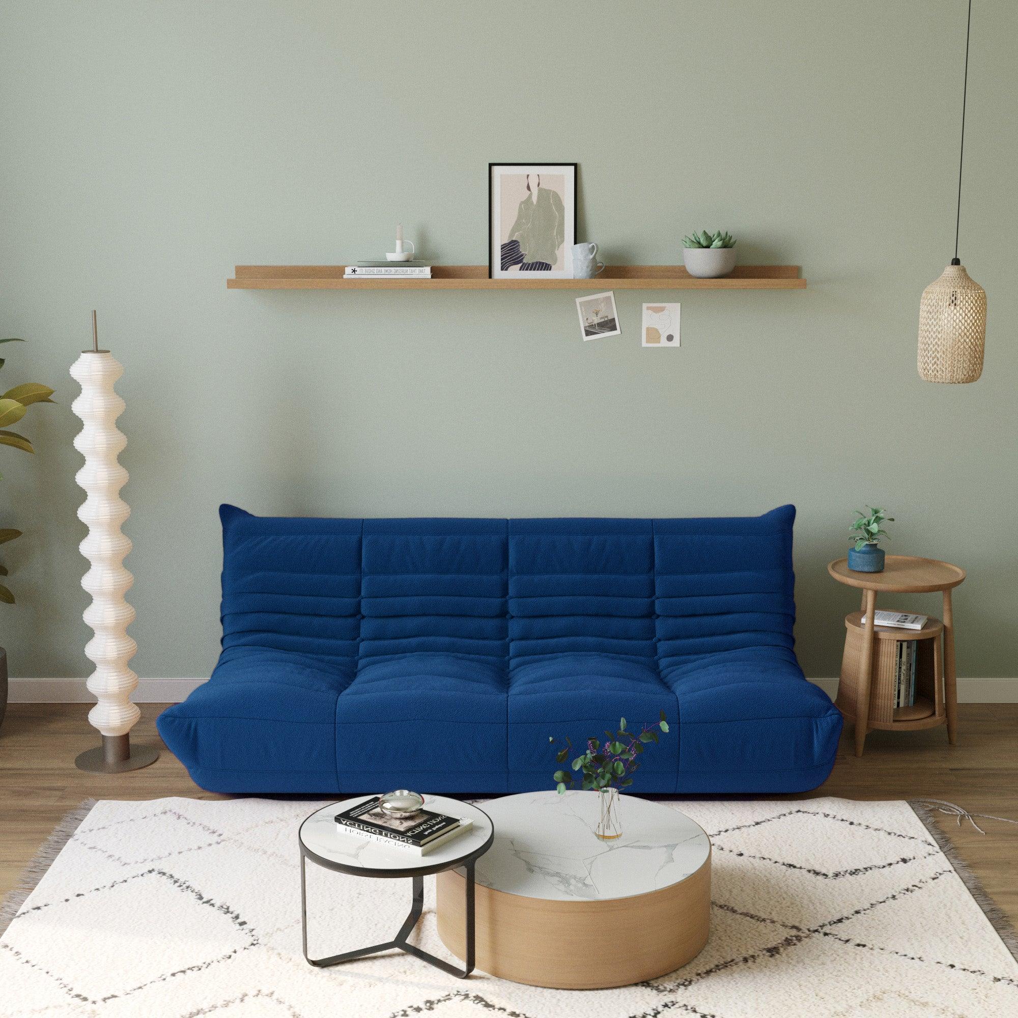 A blue, caterpillar-shaped Togo sofa with a plush texture, situated in a room with minimalist decor, on a patterned rug.