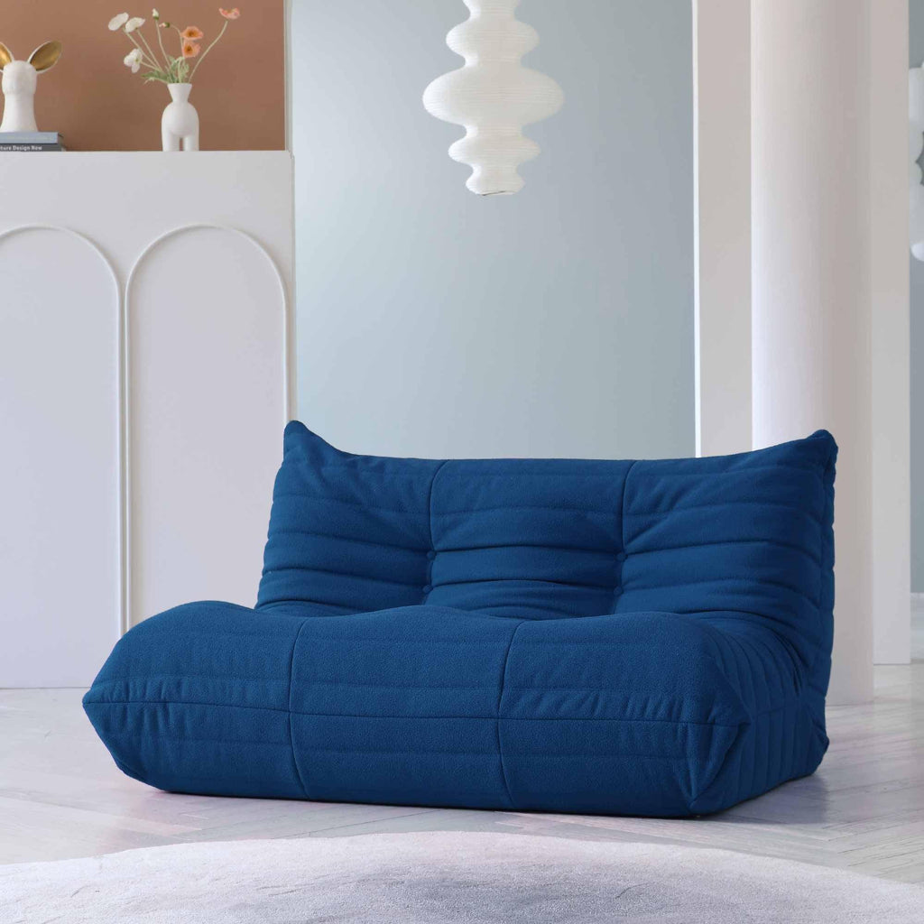 A blue, caterpillar-shaped Togo sofa on a wooden floor with a light blue wall in the background.