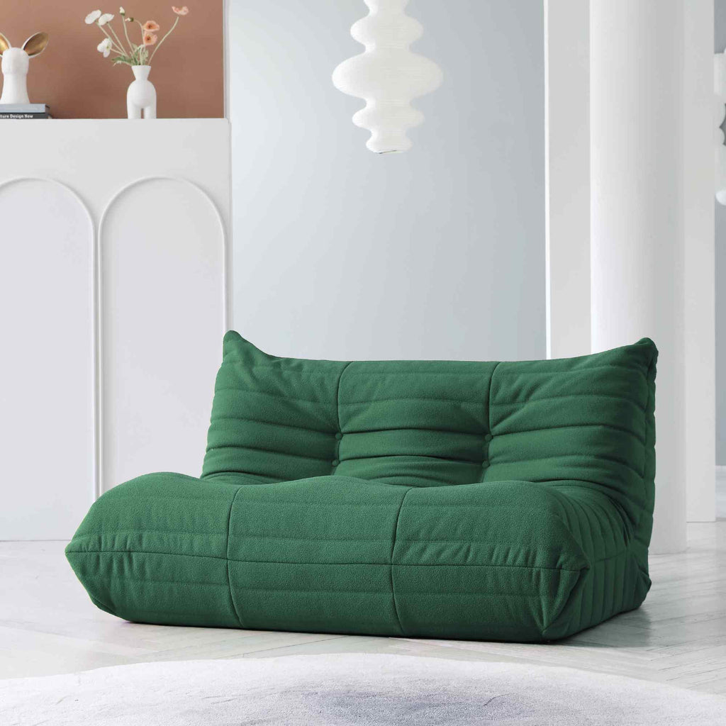 A green, caterpillar-shaped Togo sofa on a wooden floor with a light blue wall in the background.