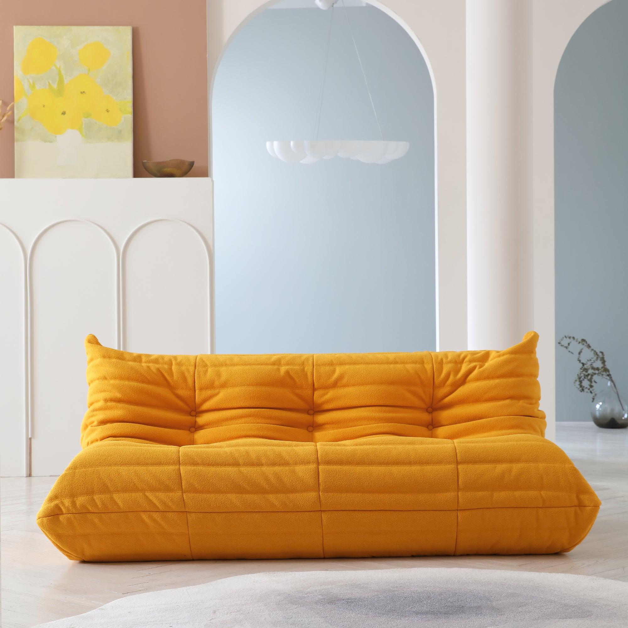 A distinctive yellow caterpillar-shaped 3-seater Togo sofa placed on a white floor in a room with light-colored walls and a painting in the background.