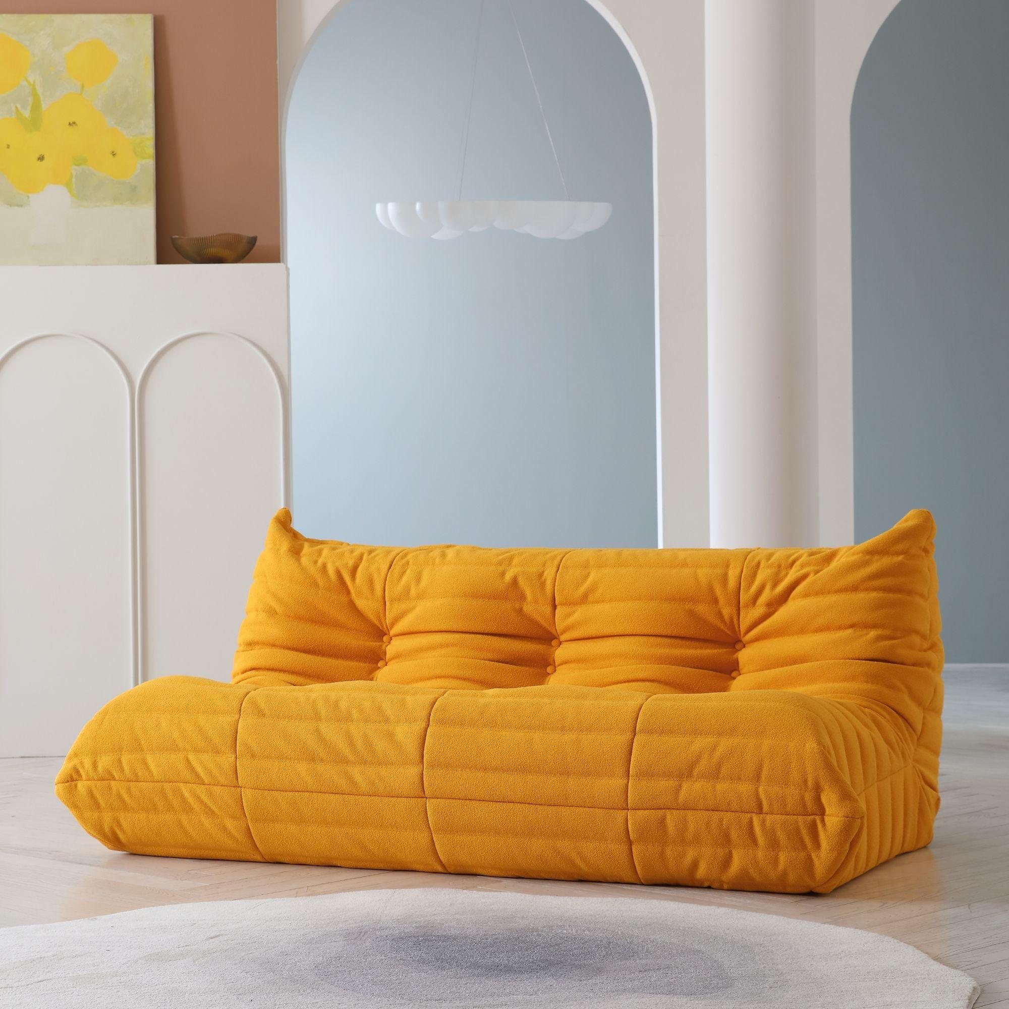 A distinctive yellow caterpillar-shaped 3-seater Togo sofa placed on a white floor in a room with light-colored walls and a painting in the background.