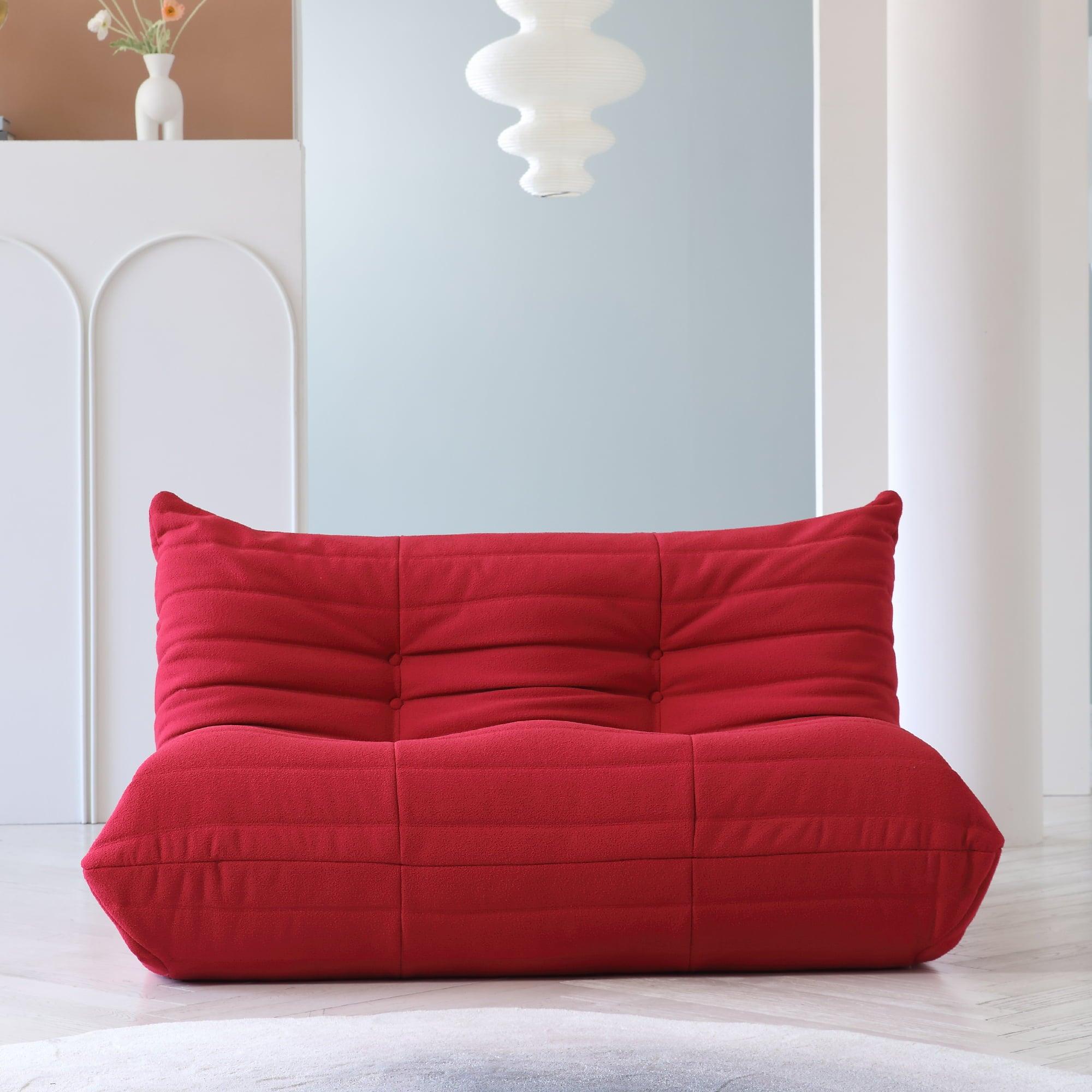 A red, caterpillar-shaped Togo sofa on a wooden floor with a light blue wall in the background.