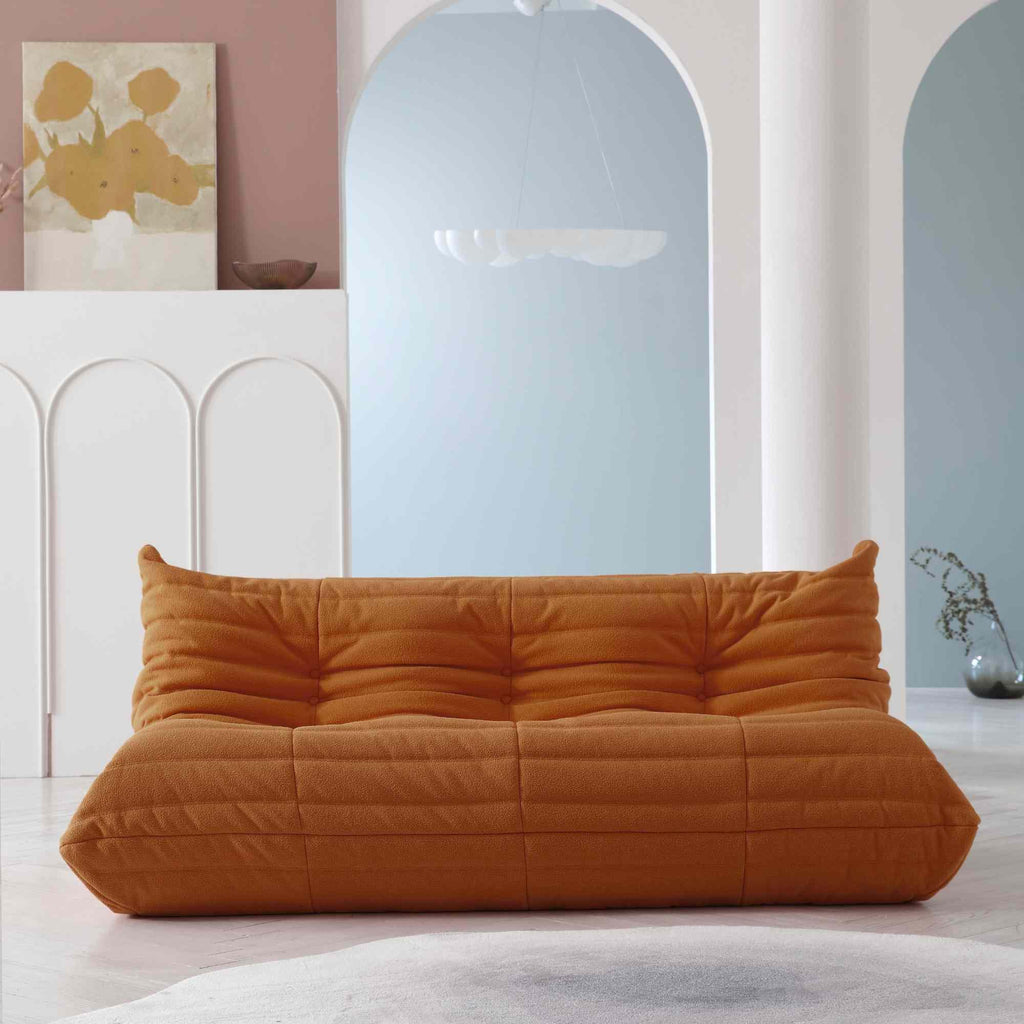 A distinctive brown caterpillar-shaped 3-seater Togo sofa placed on a white floor in a room with light-colored walls and a painting in the background.
