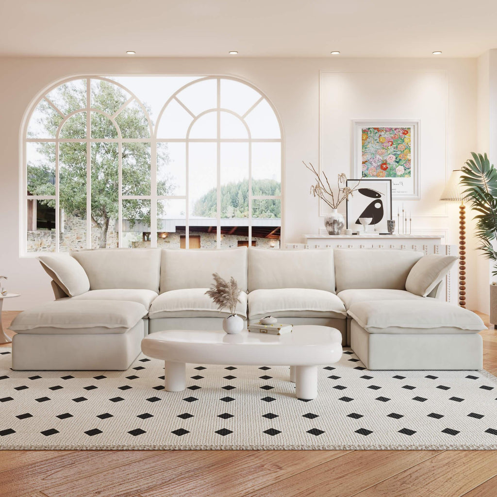 A beige U-shaped linen 6-seat sectional sofa with a storage ottoman situated in a bright room with large windows, alongside a low coffee table with items on it.