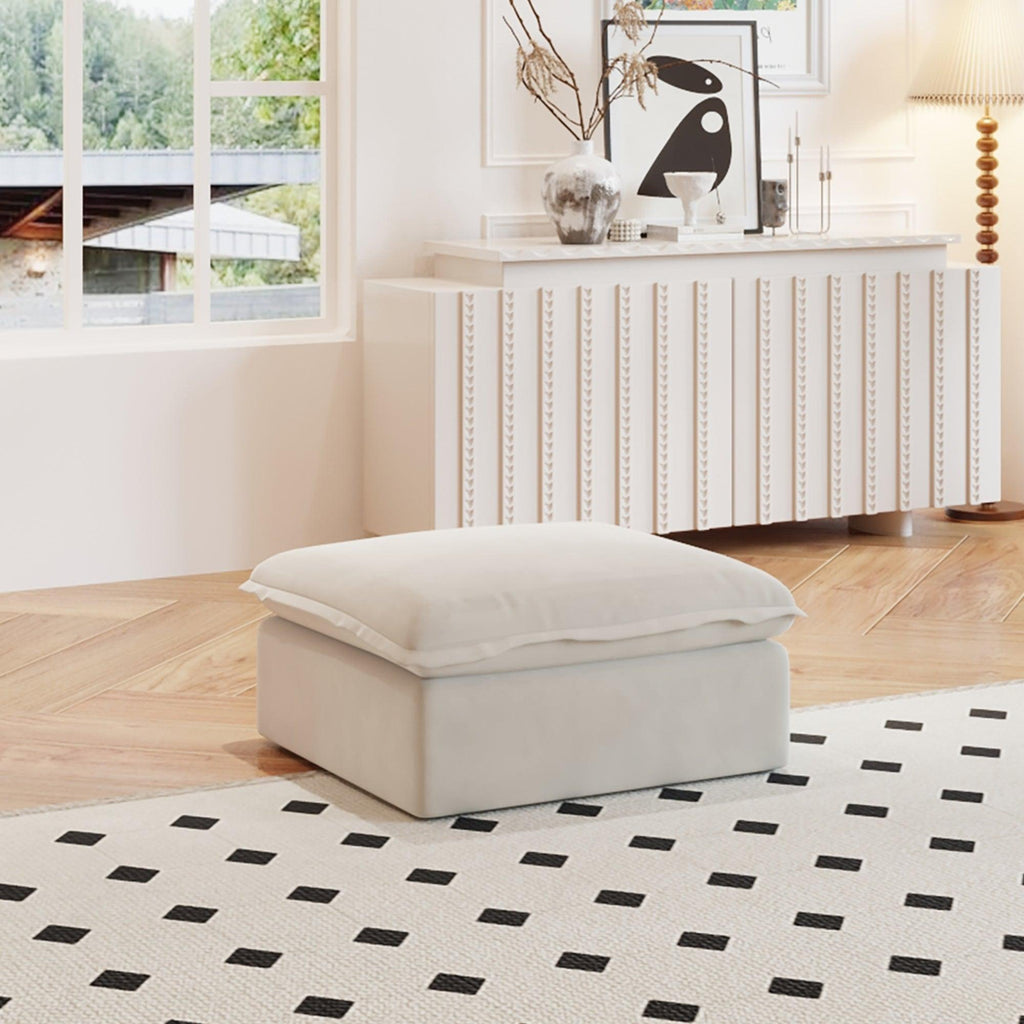 A beige linen ottoman with a cushion on top, placed on a black and white patterned rug in a room with a window and a cozy interior decor.