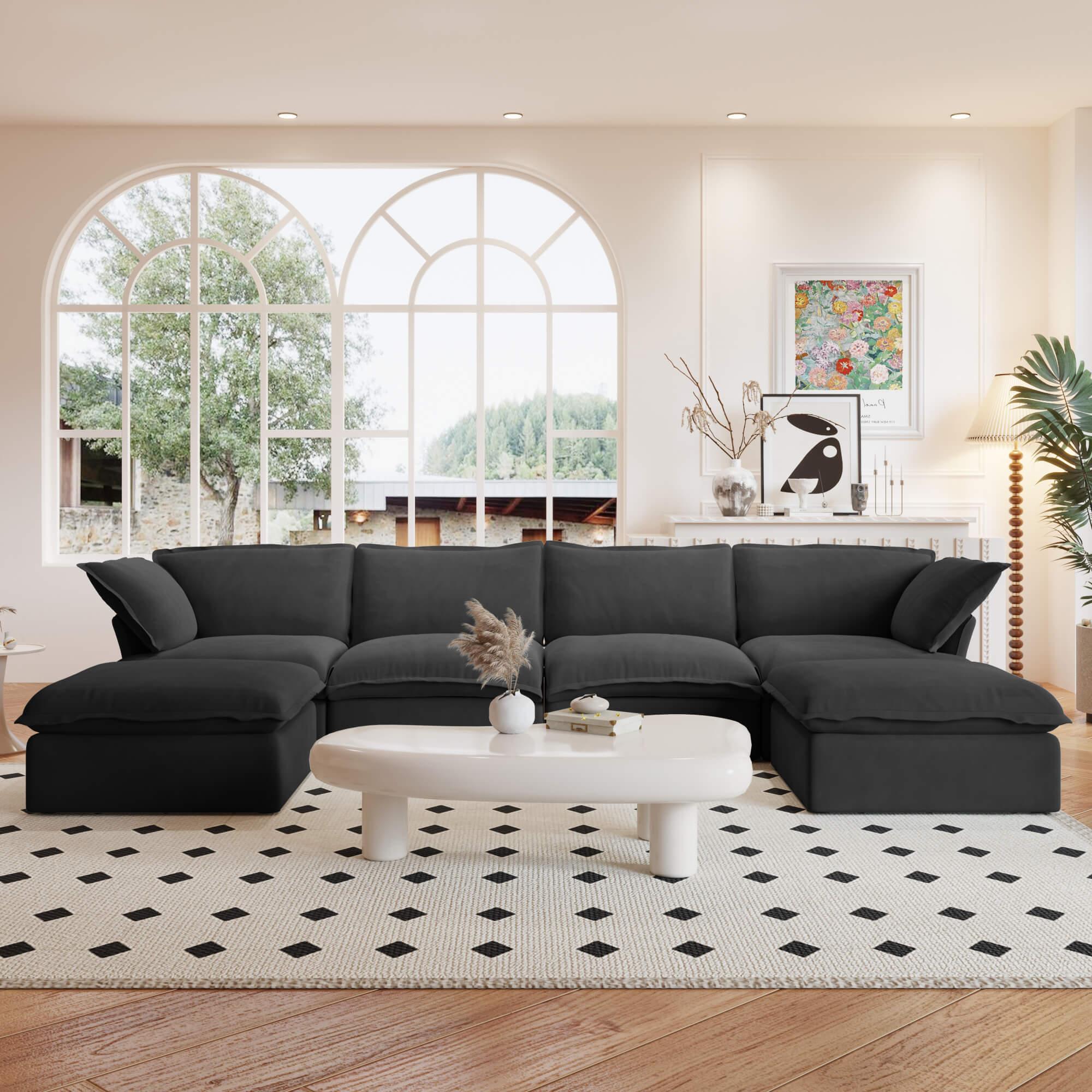 A black U-shaped linen 6-seat sectional sofa with a storage ottoman situated in a bright room with large windows, alongside a low coffee table with items on it.