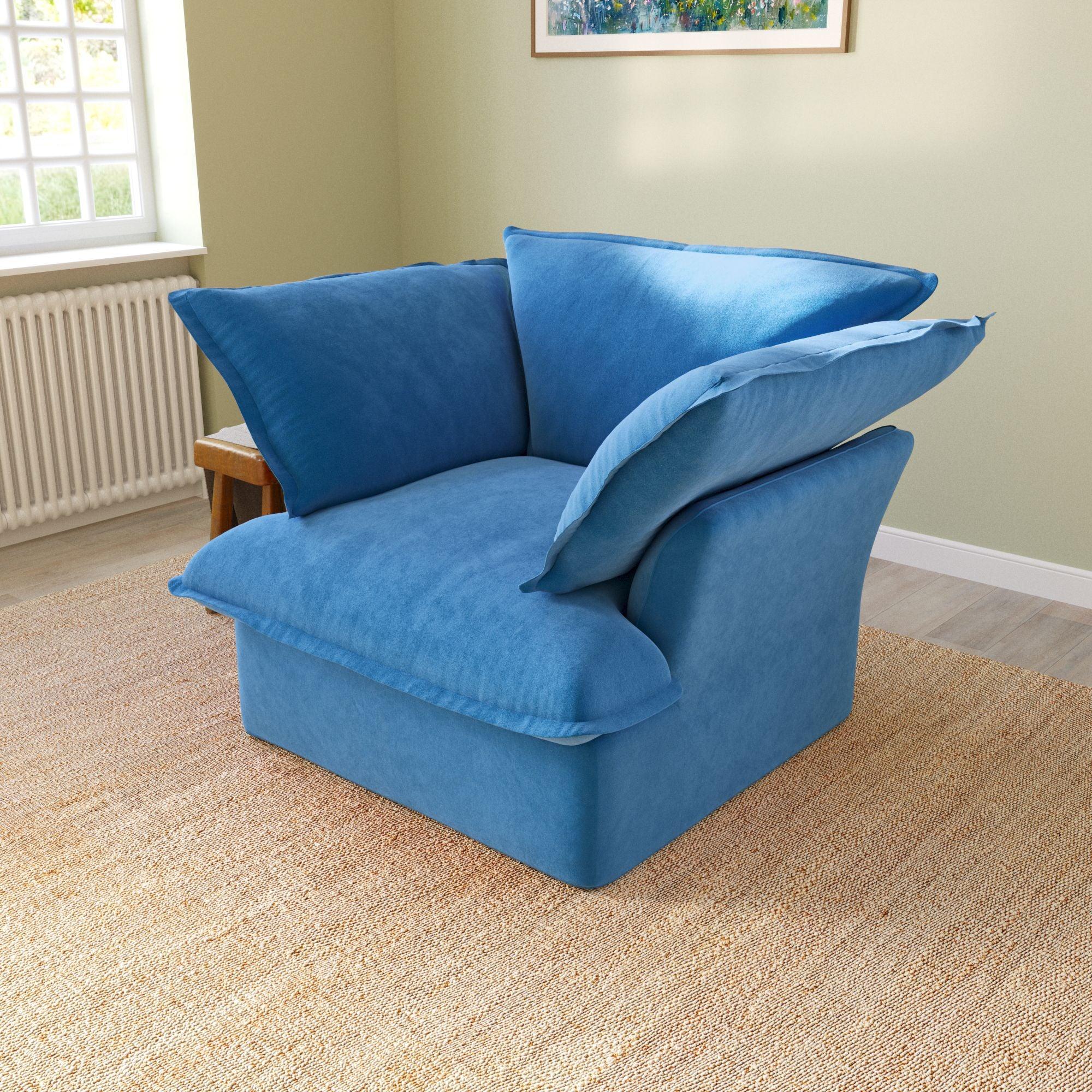 A blue velvet accent chair with a cushioned backrest and seat, placed in a room with a light green wall and a framed picture on the wall.
