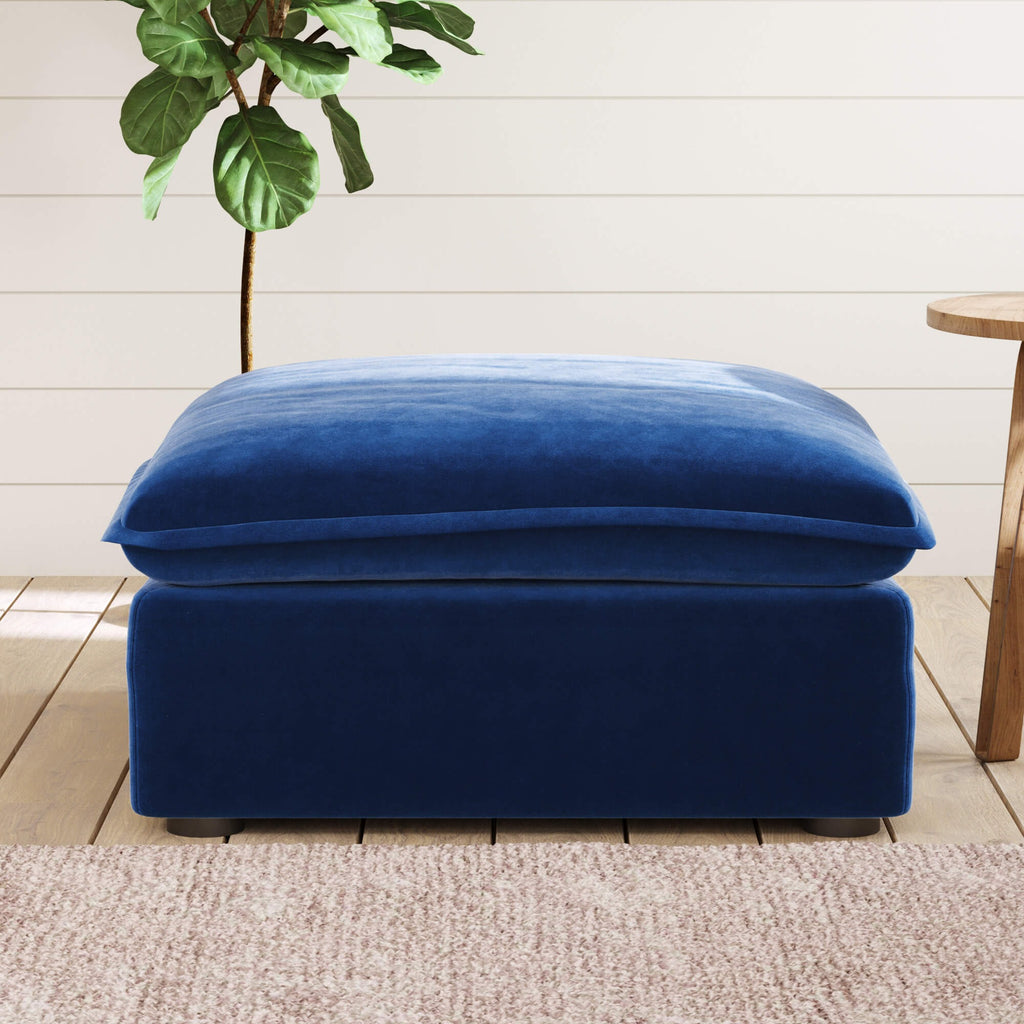Dark blue ottoman with a cushion on a wooden floor with a plant in the background