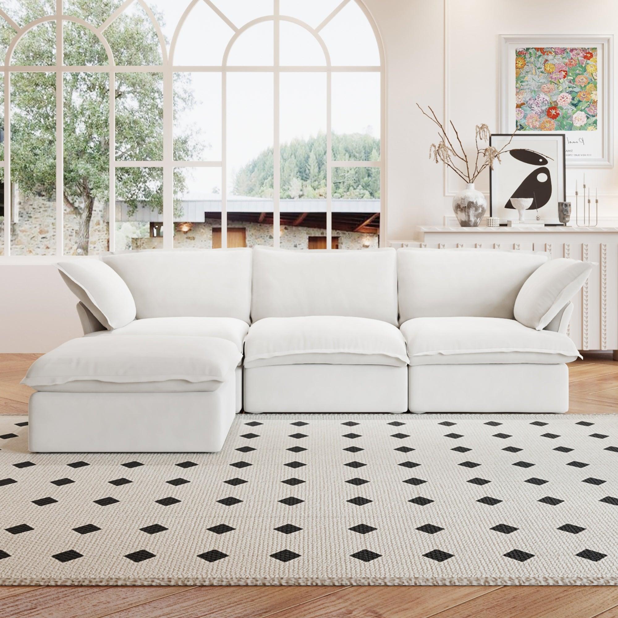 A white L-shaped linen sectional sofa with a storage ottoman positioned in a bright room with large windows, alongside a patterned rug and in front of a wall with artwork.