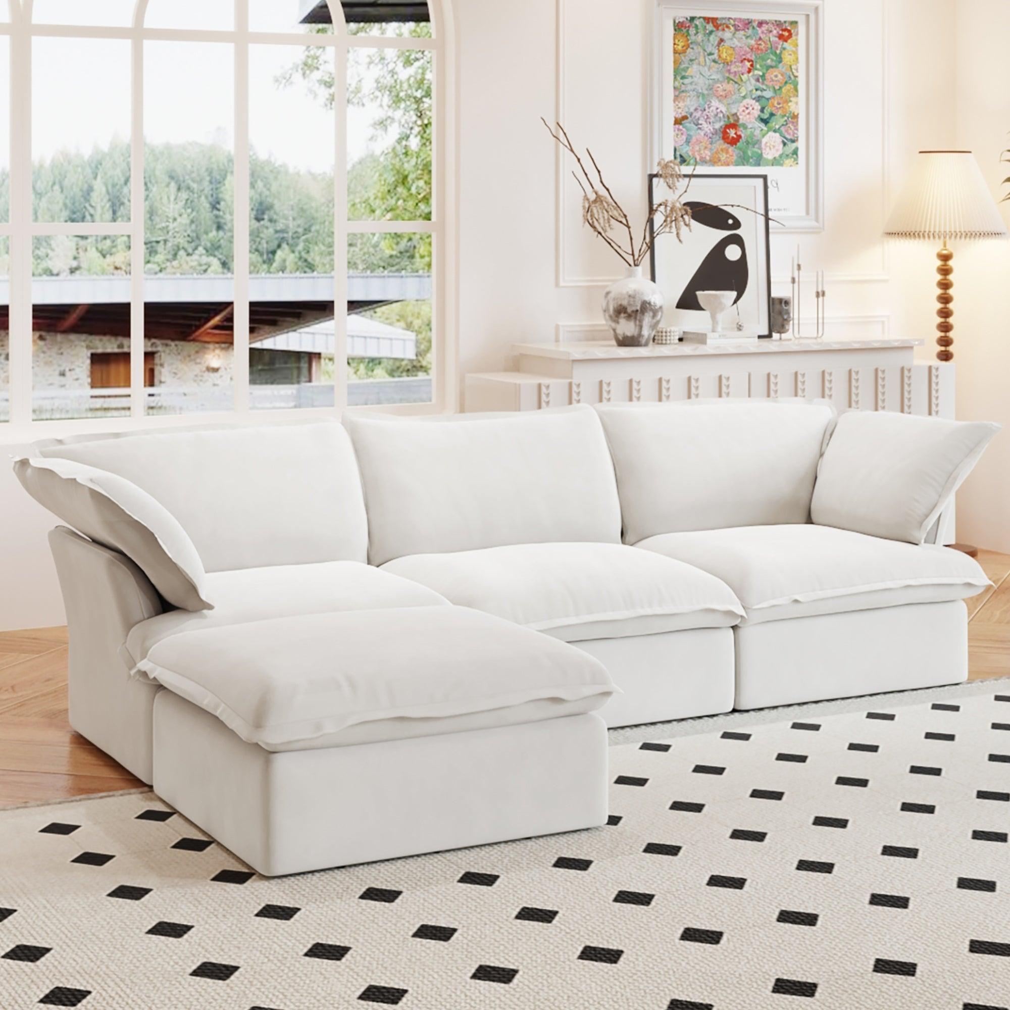 A white L-shaped linen sectional sofa with a storage ottoman positioned in a bright room with large windows, alongside a patterned rug and in front of a wall with artwork.