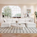 A white U-shaped linen sectional sofa with a storage ottoman situated in a bright room with large windows, alongside a low coffee table with items on it.