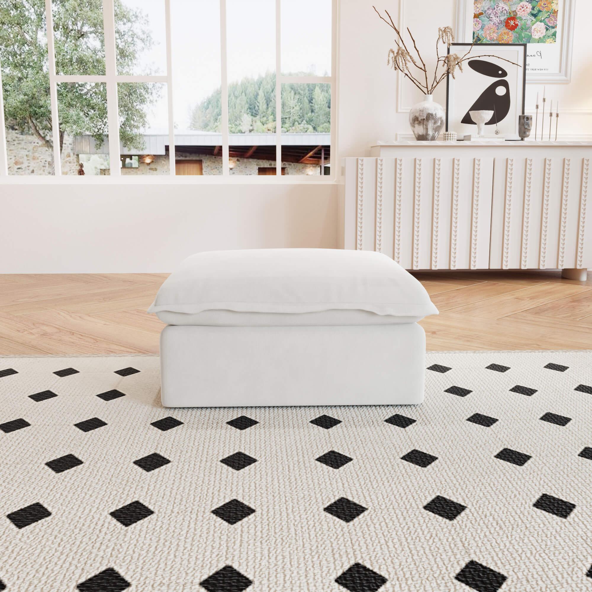 A white linen sectional ottoman with storage compartment placed on a black and white patterned rug in a living room setting.