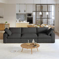 A modern black 3-seater sectional sofa in a living room setting with a round wooden coffee table in front and a kitchen area in the background.