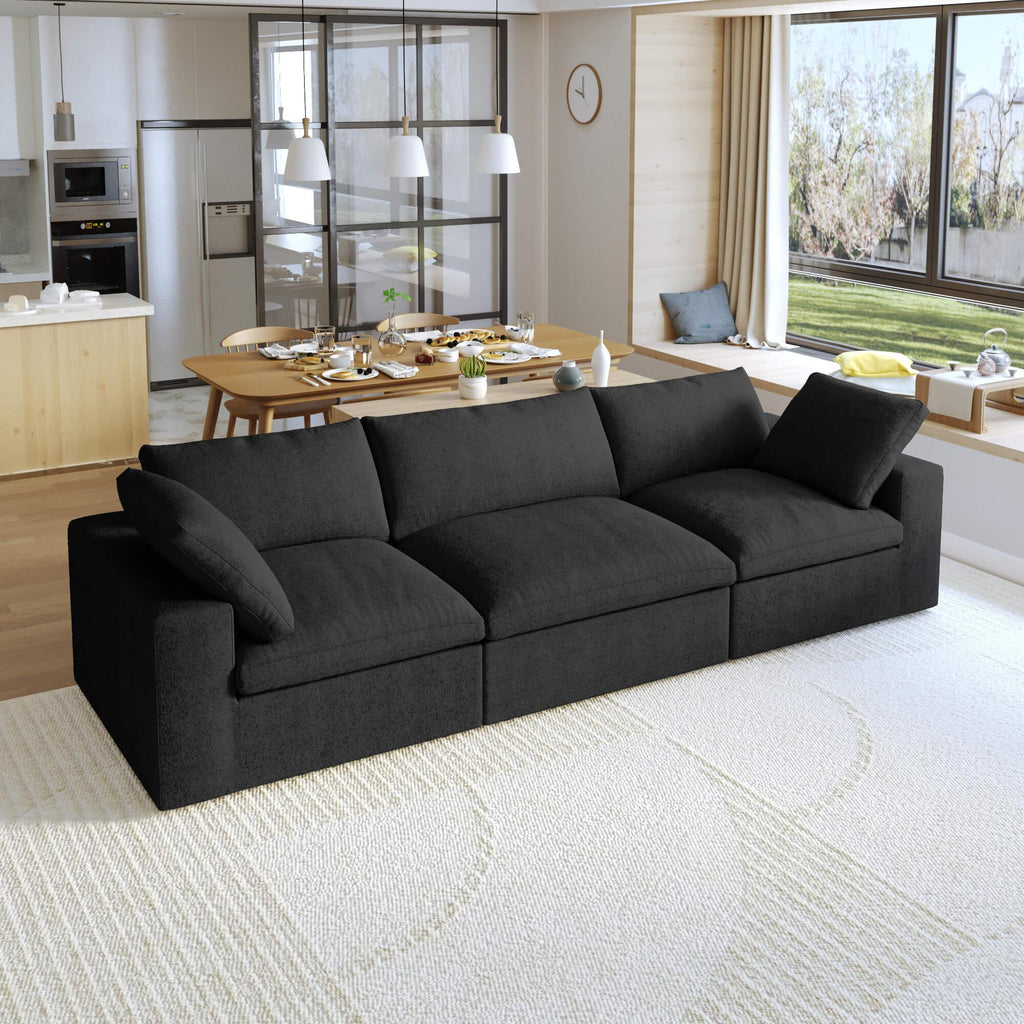 A modern black 3-seater sectional sofa in a living room setting with a round wooden coffee table in front and a kitchen area in the background.