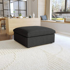 A black, down-filled comfort storage ottoman, placed in a bright living room setting.