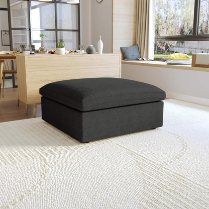 A black, down-filled comfort storage ottoman, placed in a bright living room setting.