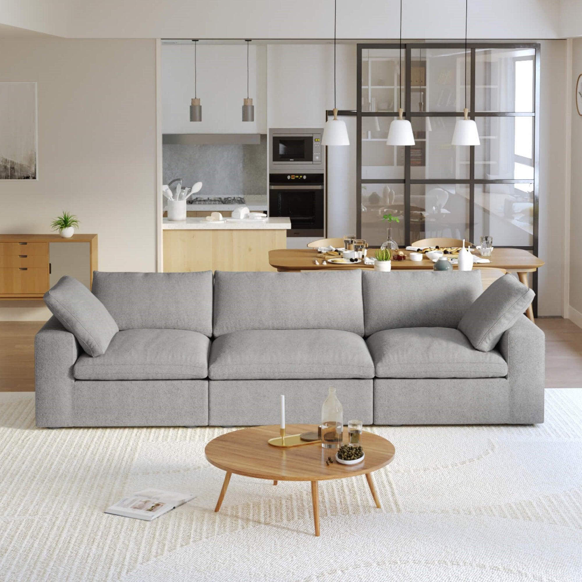 A modern gray 3-seater sectional sofa in a living room setting with a round wooden coffee table in front and a kitchen area in the background.