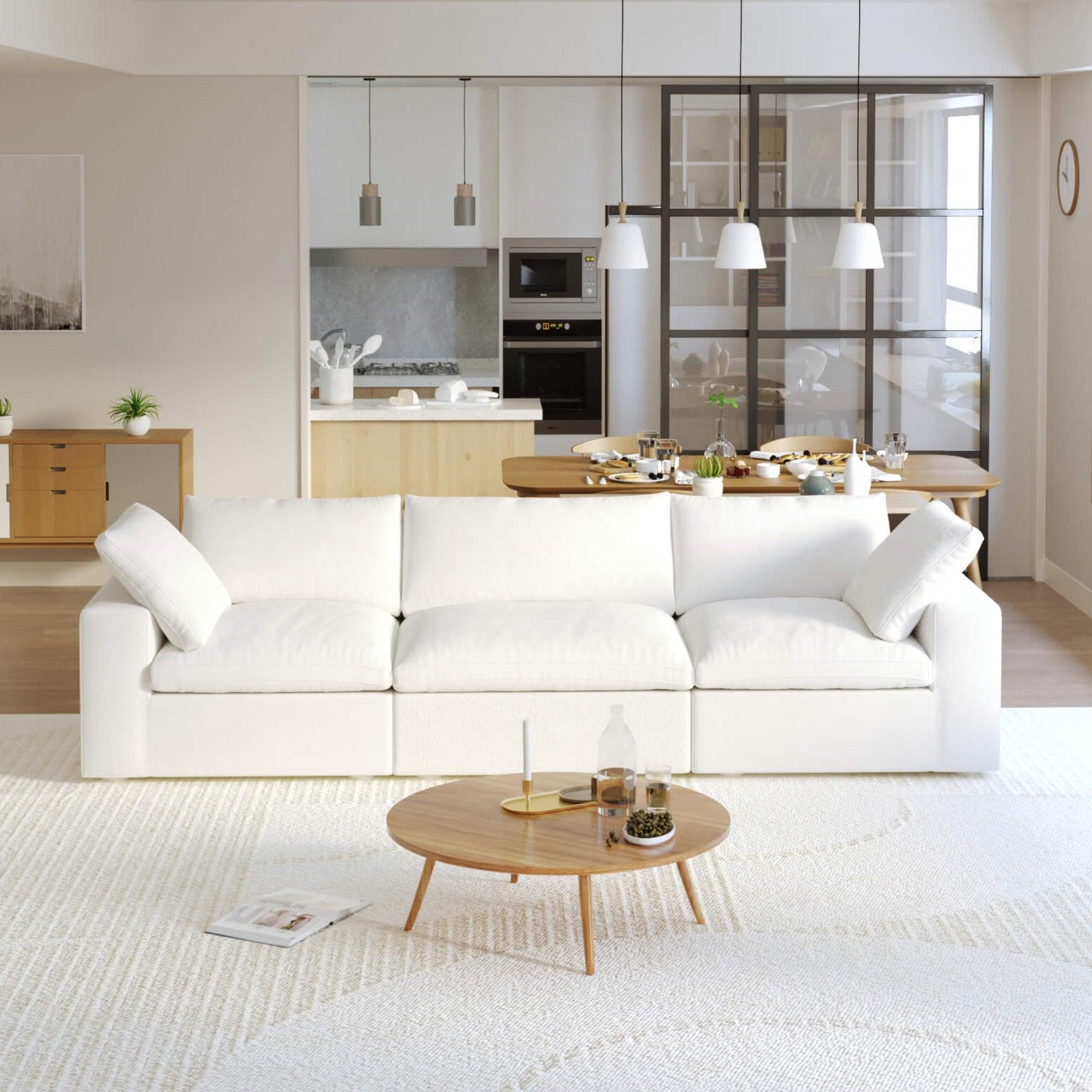 A modern white 3-seater sectional sofa in a living room setting with a round wooden coffee table in front and a kitchen area in the background.