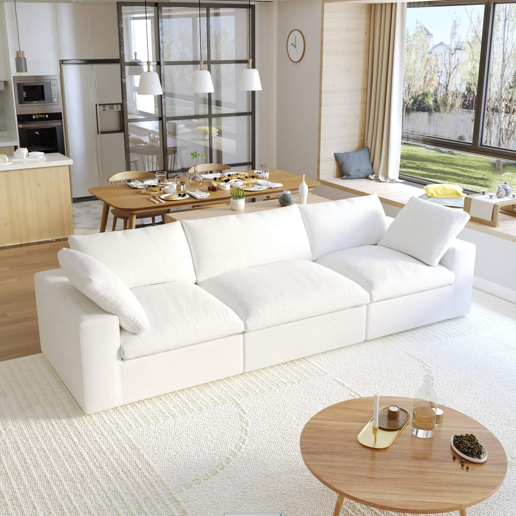 A modern white 3-seater sectional sofa in a living room setting with a round wooden coffee table in front and a kitchen area in the background.