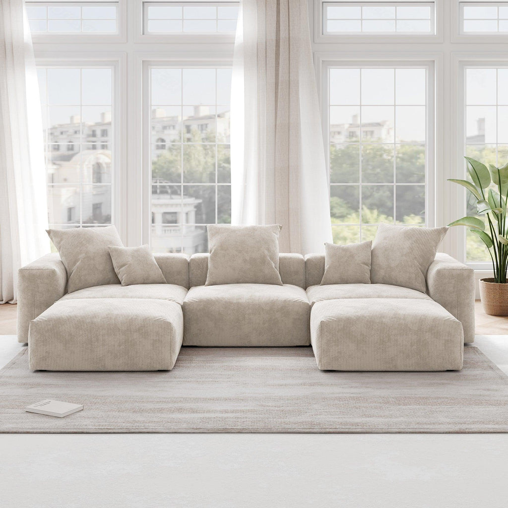A beige corduroy U-shaped sectional modular sofa with detachable armrests and removable covers, placed in a room with large windows.