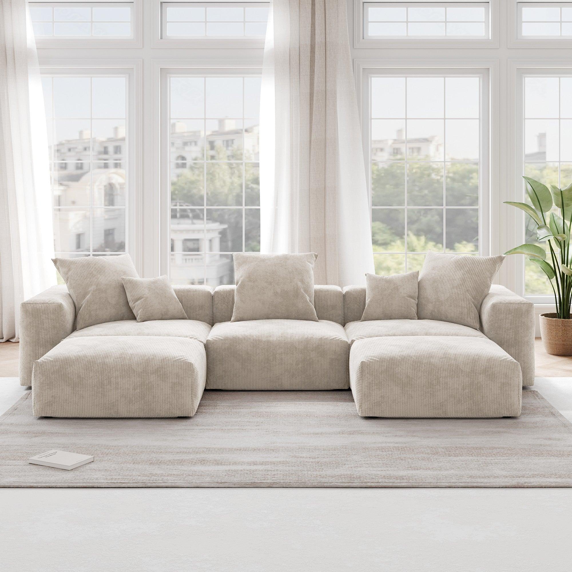 A beige corduroy U-shaped sectional modular sofa with detachable armrests and removable covers, placed in a room with large windows.