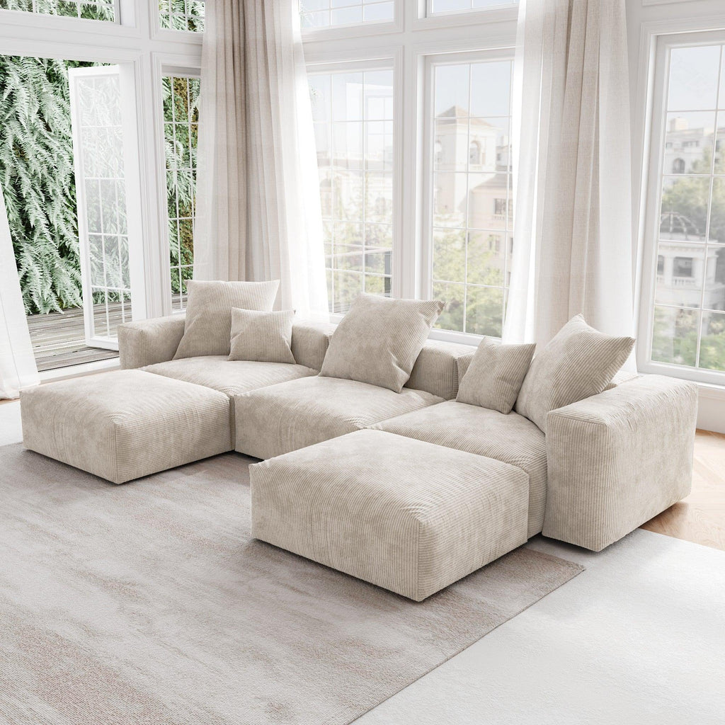 A beige corduroy U-shaped sectional modular sofa with detachable armrests and removable covers, placed in a room with large windows.