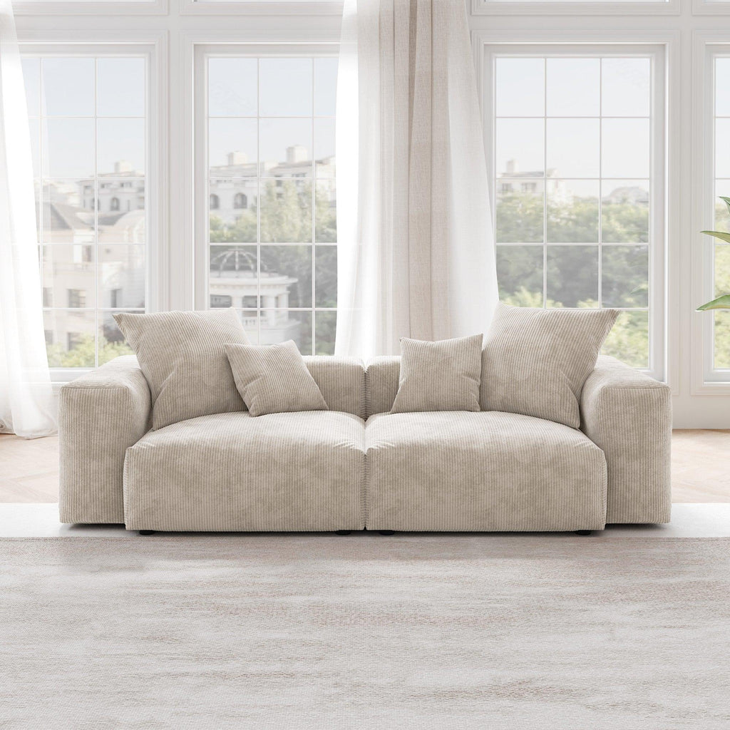 A beige corduroy sectional loveseat sofa with detachable armrests and removable covers, positioned in a bright room with large windows.