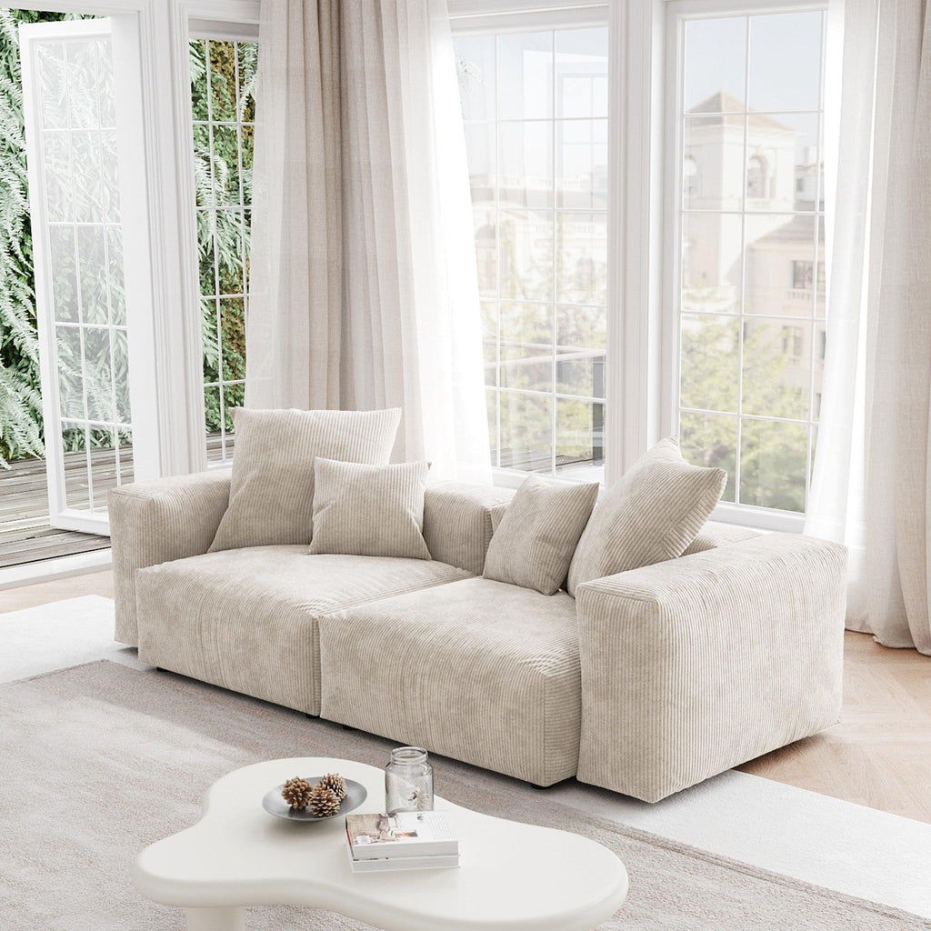 A beige corduroy sectional loveseat sofa with detachable armrests and removable covers, positioned in a bright room with large windows.