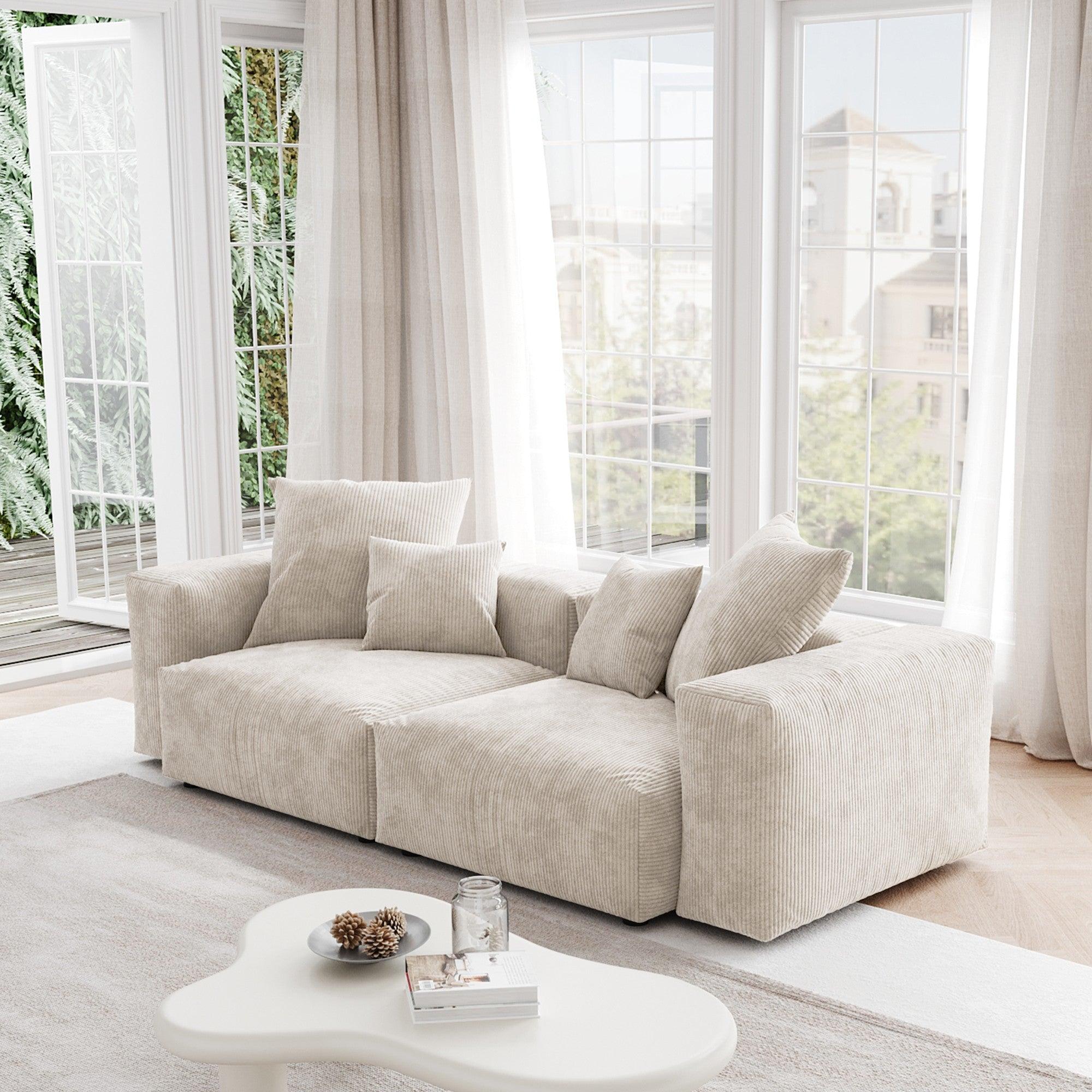 A beige corduroy sectional loveseat sofa with detachable armrests and removable covers, positioned in a bright room with large windows.