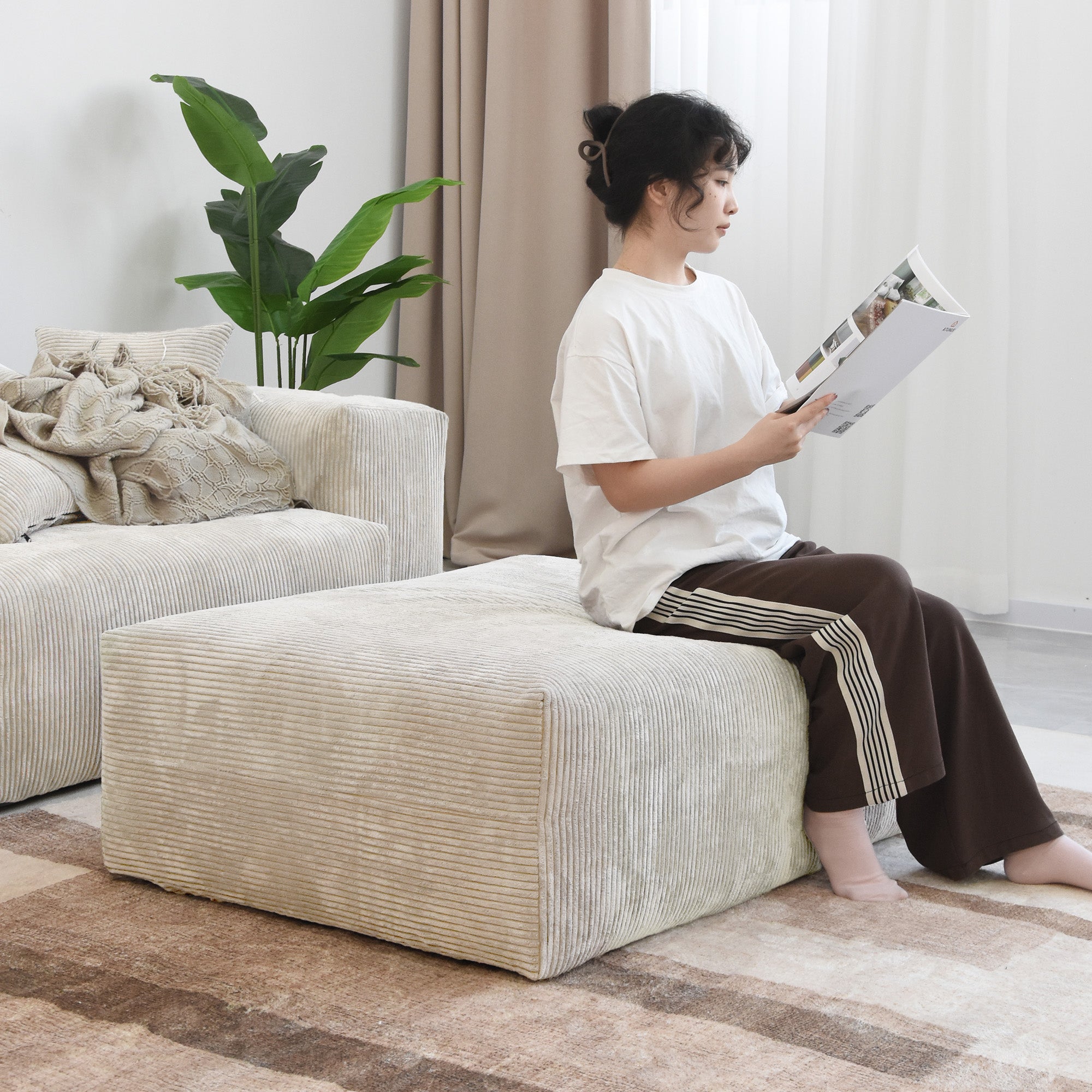 Person sitting on a beige corduroy ottoman, reading magazine in a light living room