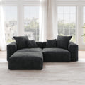 A black corduroy sectional loveseat sofa with ottoman, detachable armrests, and removable covers, featuring a chaise lounge configuration placed in a room with large windows.