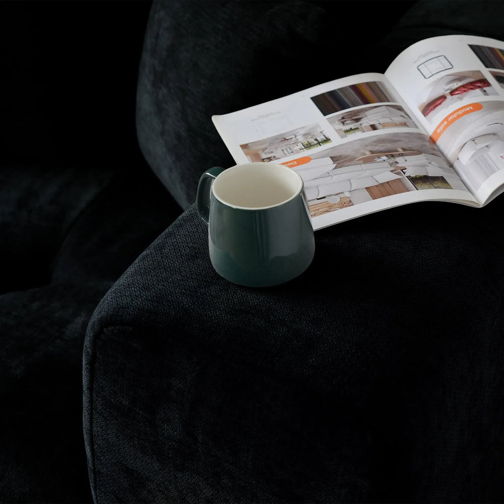 Green mug next to an open magazine on a wide armrest of a black sofa
