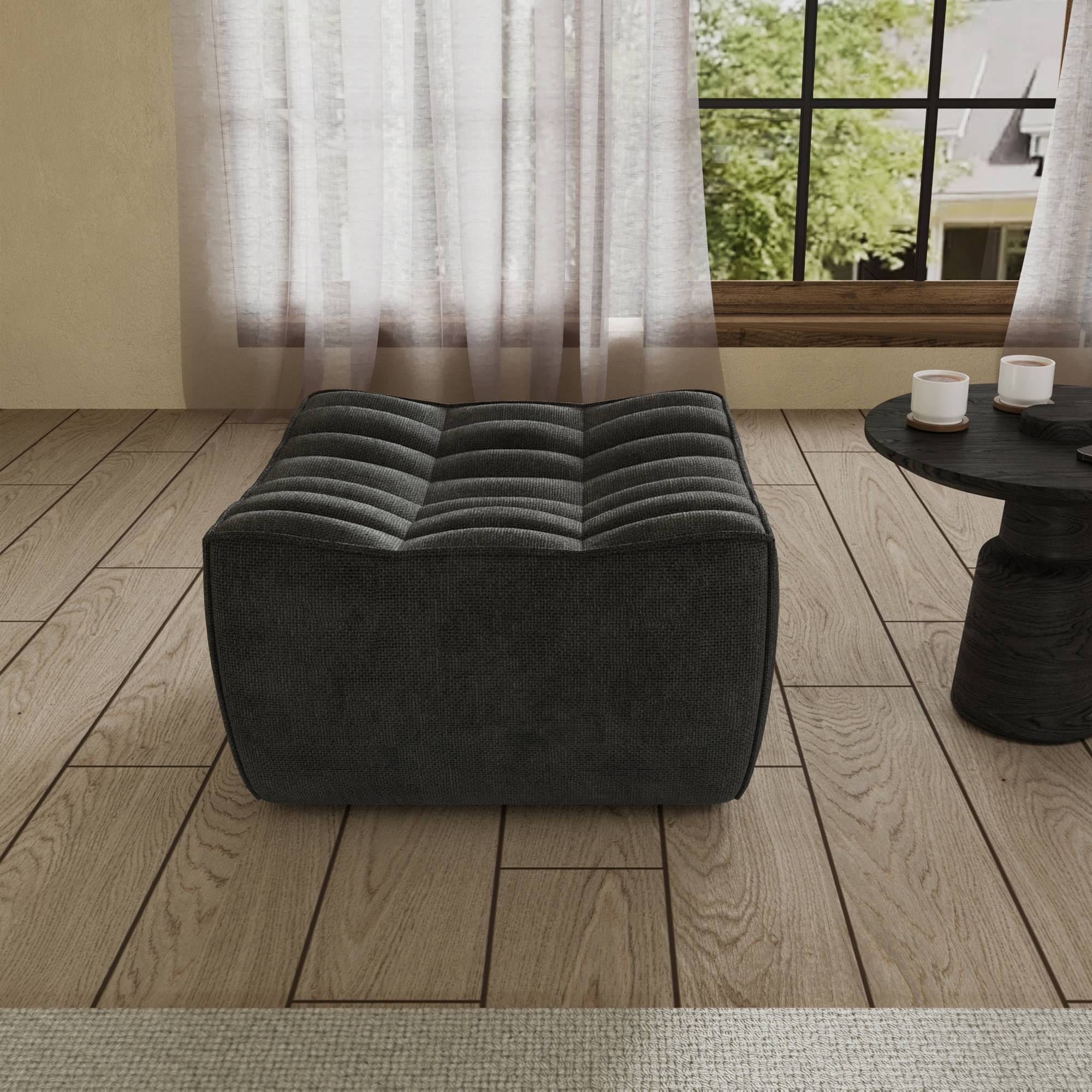 A black chenille ottoman with a minimalist design, placed on a wooden floor with a large window in the background.