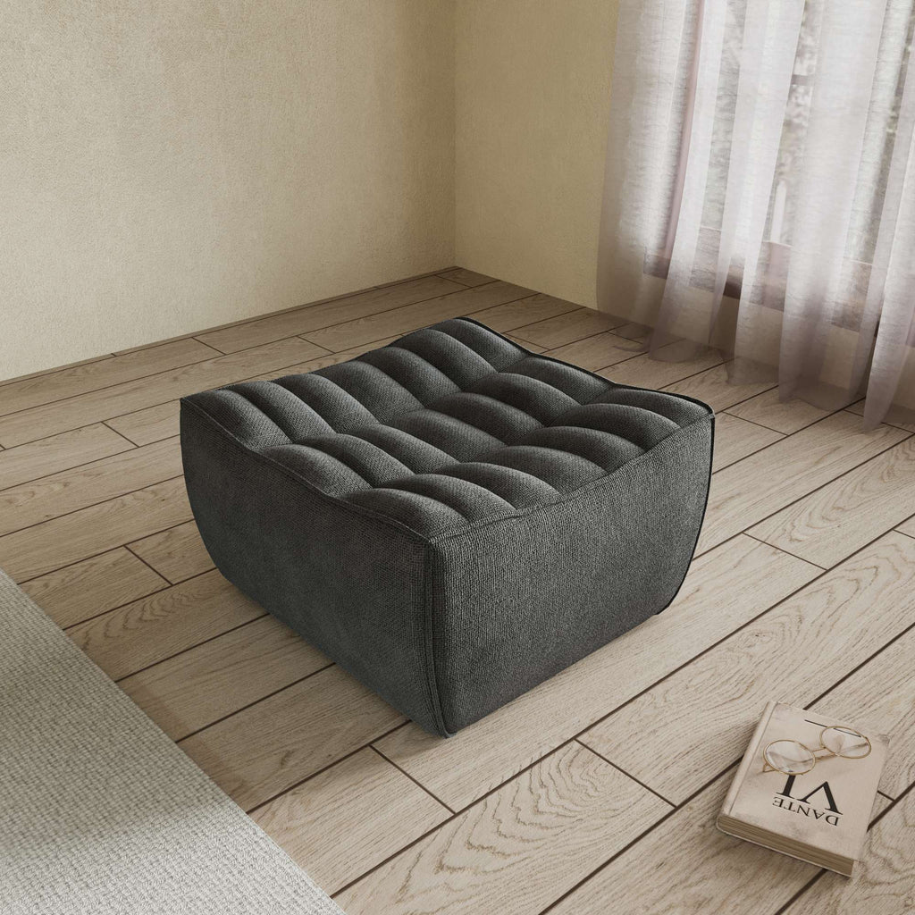 A black chenille ottoman with a minimalist design, placed on a wooden floor with a large window in the background.