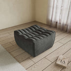 A black chenille ottoman with a minimalist design, placed on a wooden floor with a large window in the background.