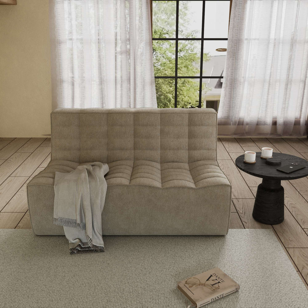 A khaki cotton-linen 2-seater ofa with an ergonomic caterpillar design, placed in a room with a window, curtains, a small round table, and a book on the floor.