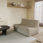 A khaki cotton-linen 2-seater ofa with an ergonomic caterpillar design, placed in a room with a window, curtains, a small round table, and a book on the floor.