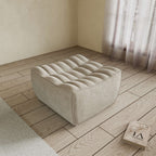 A khaki chenille ottoman with a minimalist design, placed on a wooden floor with a large window in the background.